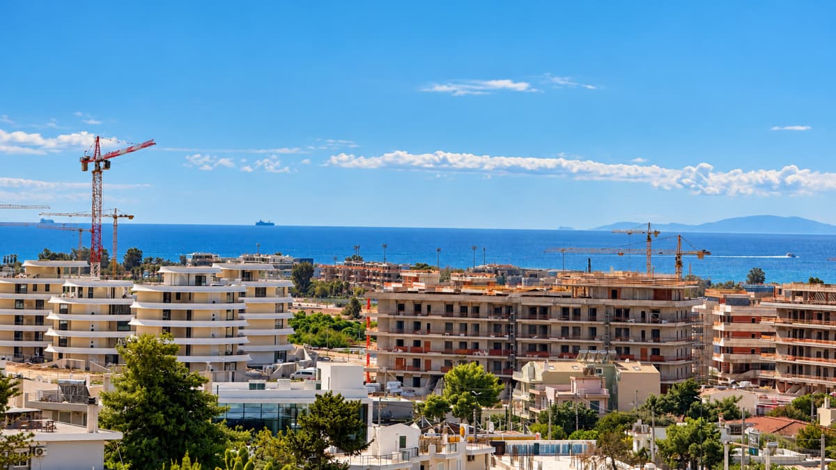 Εξωτερική άποψη εργοταξίου με πολυκατοικίες υπό κατασκευή, γερανούς και θάλασσα στο βάθος.