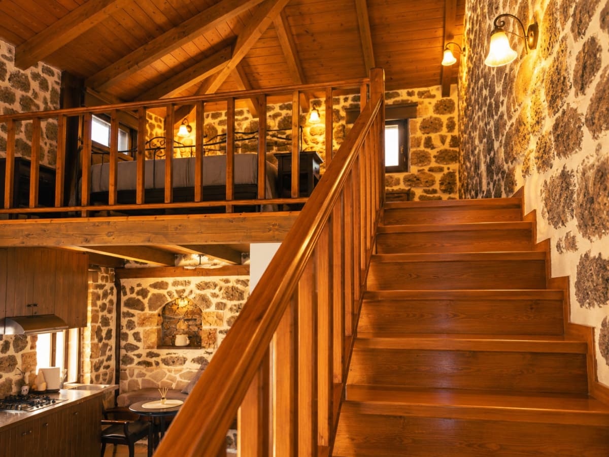 A wooden staircase with a banister leads upward beside a stone wall, with a loft bed visible on the second level above a small kitchen.