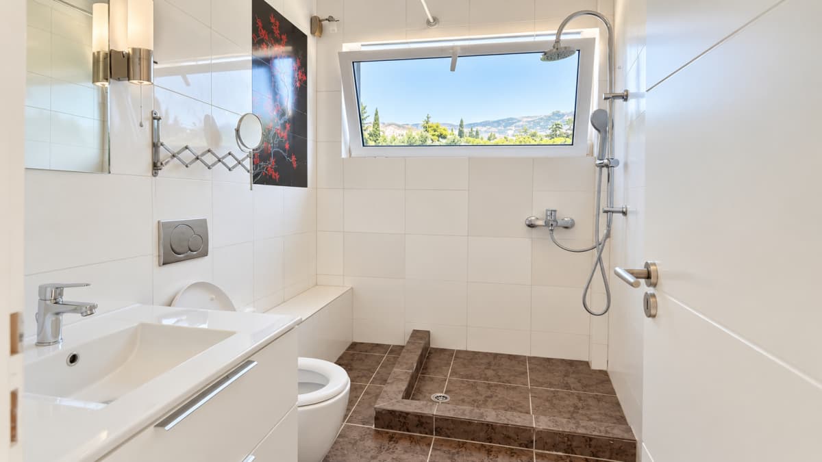 Bathroom with a raised tiled shower area, a white vanity and a window above the shower.