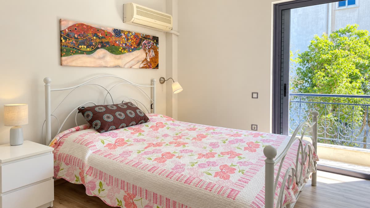 Bedroom with a white iron bed, a painting above it and a glass door to a balcony with trees.