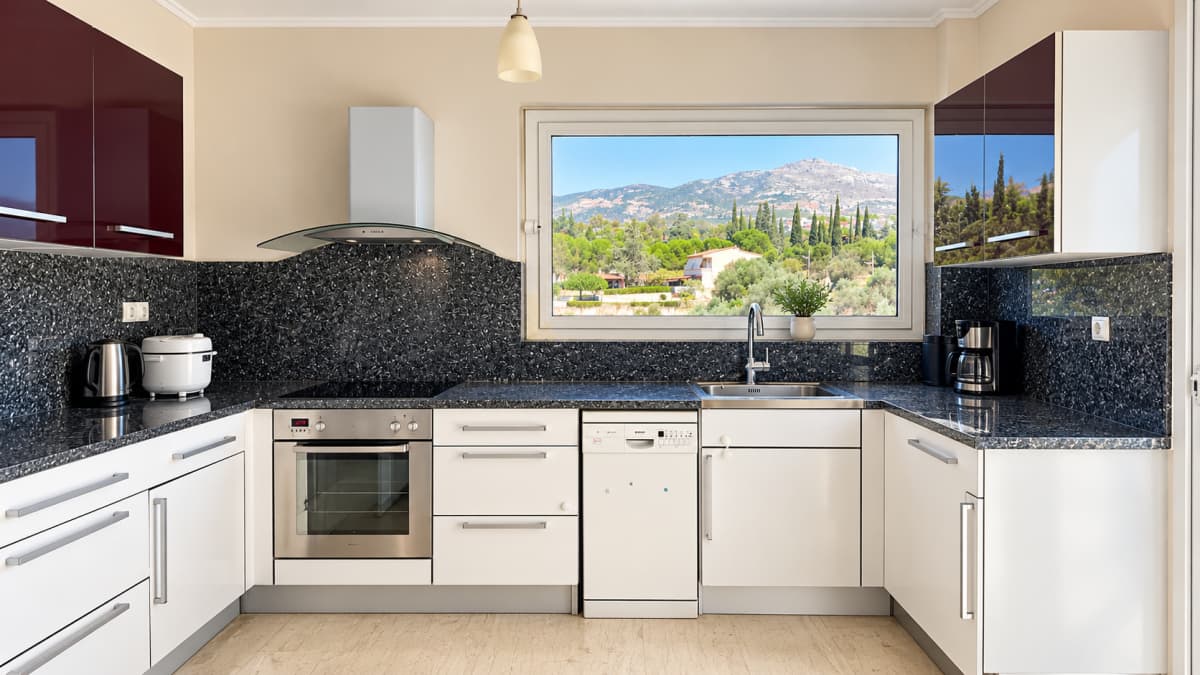 Kitchen with white and burgundy units, a dark stone worktop, built-in oven and a window onto greenery.