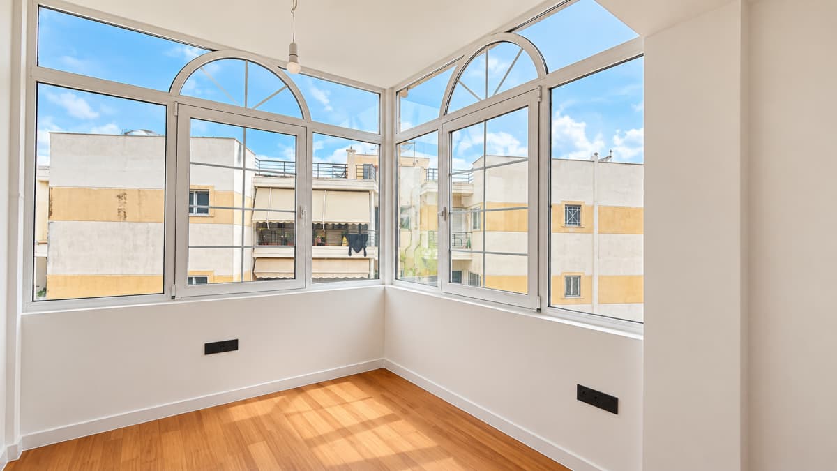 An empty room with light wood flooring, white walls, two black electrical outlets, and large arched windows overlooking buildings.