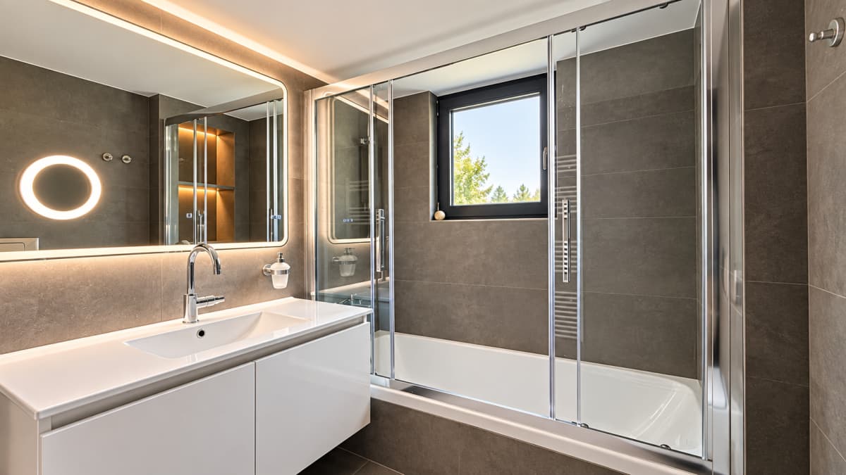 A modern bathroom featuring a white vanity with a basin sink, an illuminated mirror, and a bathtub enclosed by glass doors.