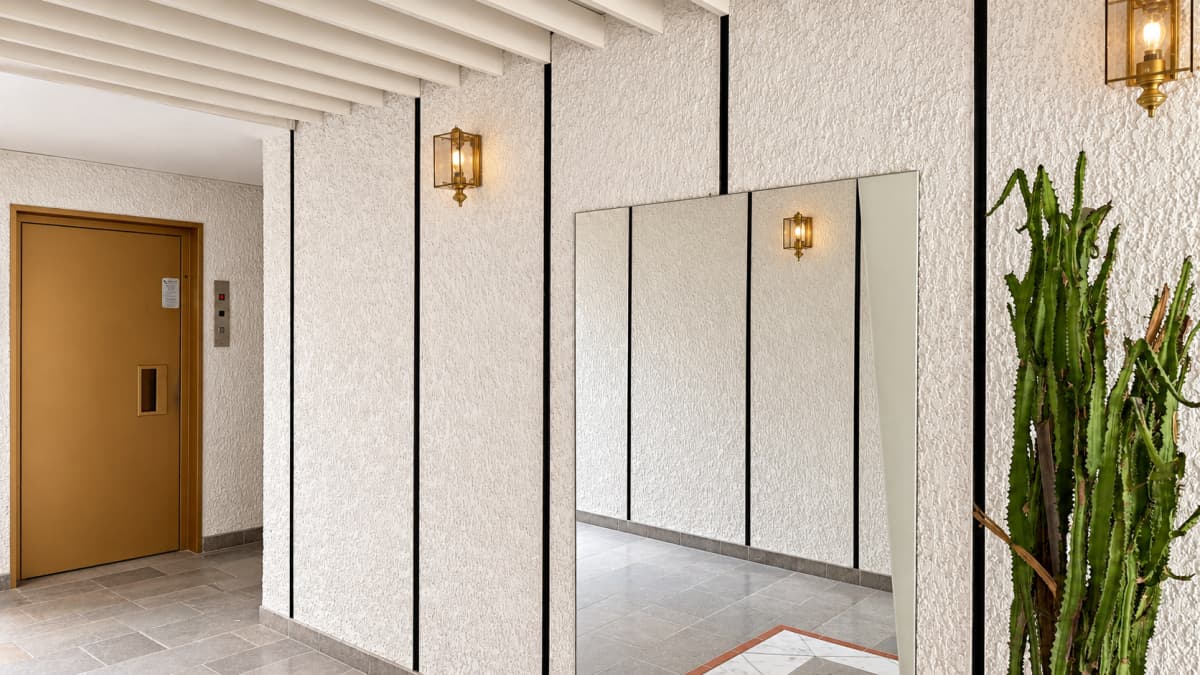 An indoor hallway with textured white walls, vertical black lines, wall sconces, mirrors, and a cactus on the right.