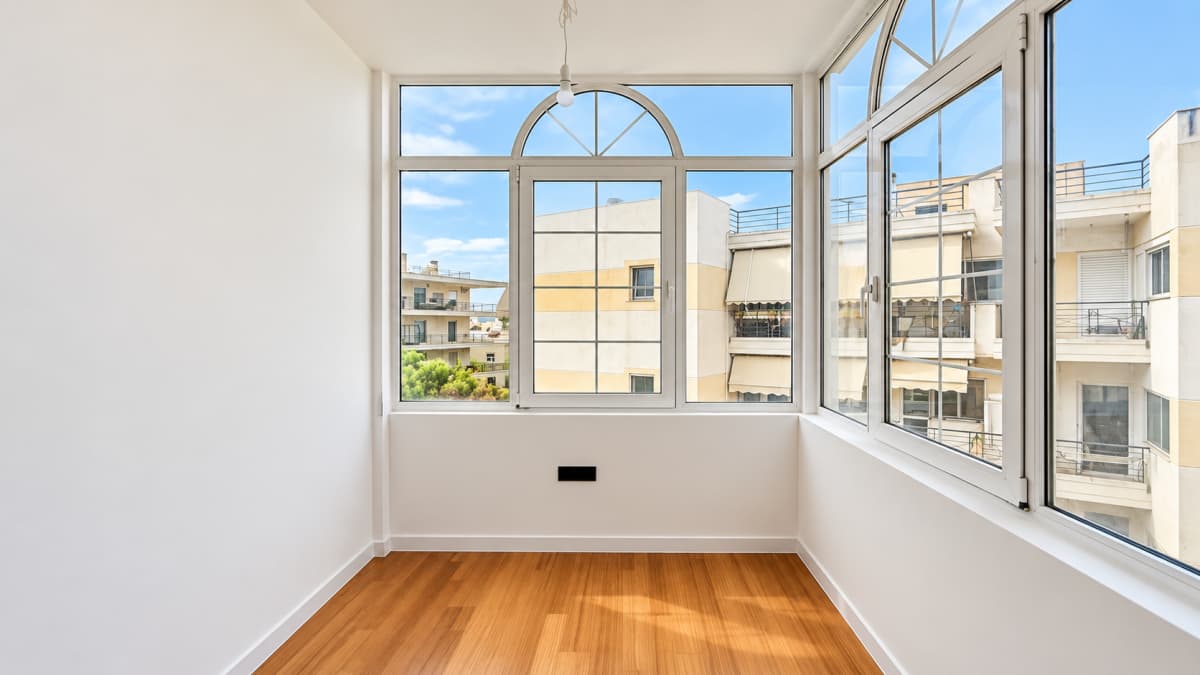 An empty enclosed balcony with white walls, a wooden floor, and large windows overlooking an apartment building.