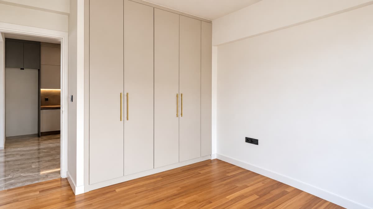 A minimal bedroom interior features light gray built-in wardrobes with vertical gold handles on a wooden floor and white walls.