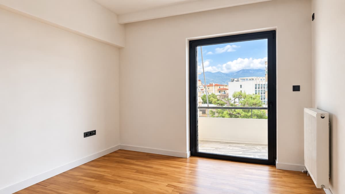 An empty room with white walls, wood flooring, a white radiator, and a large black-framed balcony door showing an outdoor urban view.
