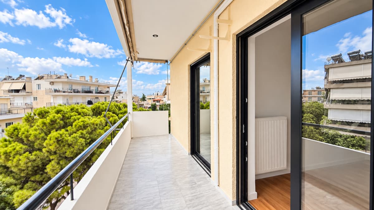 Outdoor balcony with grey tile flooring, a white railing, and a sliding glass door, overlooking trees and buildings.