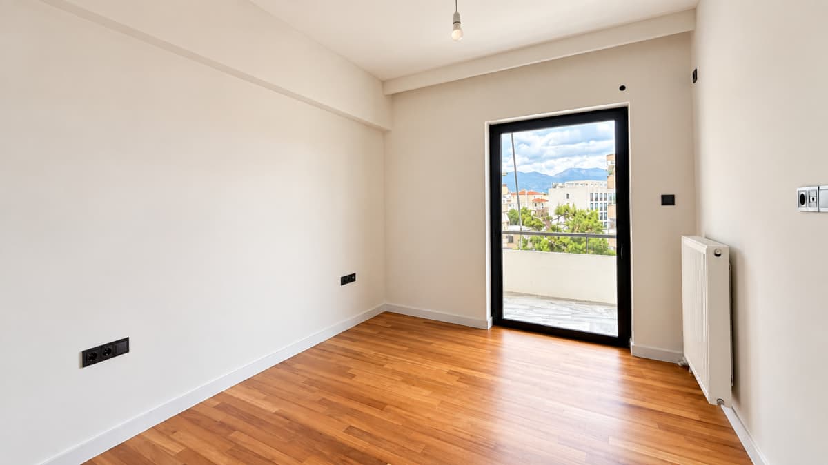 An empty room with white walls, polished wooden floors, a radiator on the right wall, and a black-framed glass door leading to a balcony.