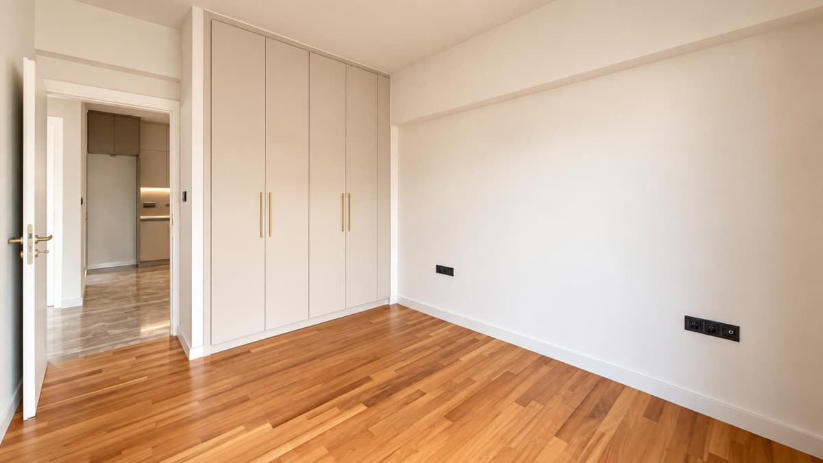An empty bedroom with light-colored walls, wood flooring, built-in wardrobes, and an open doorway.