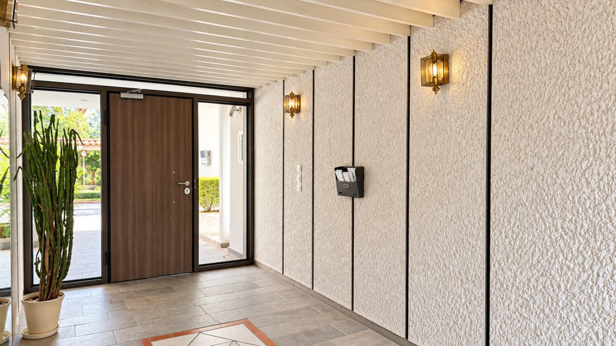 A modern building entrance hallway featuring textured off-white walls, a wooden front door with sidelights, and gray tile flooring.