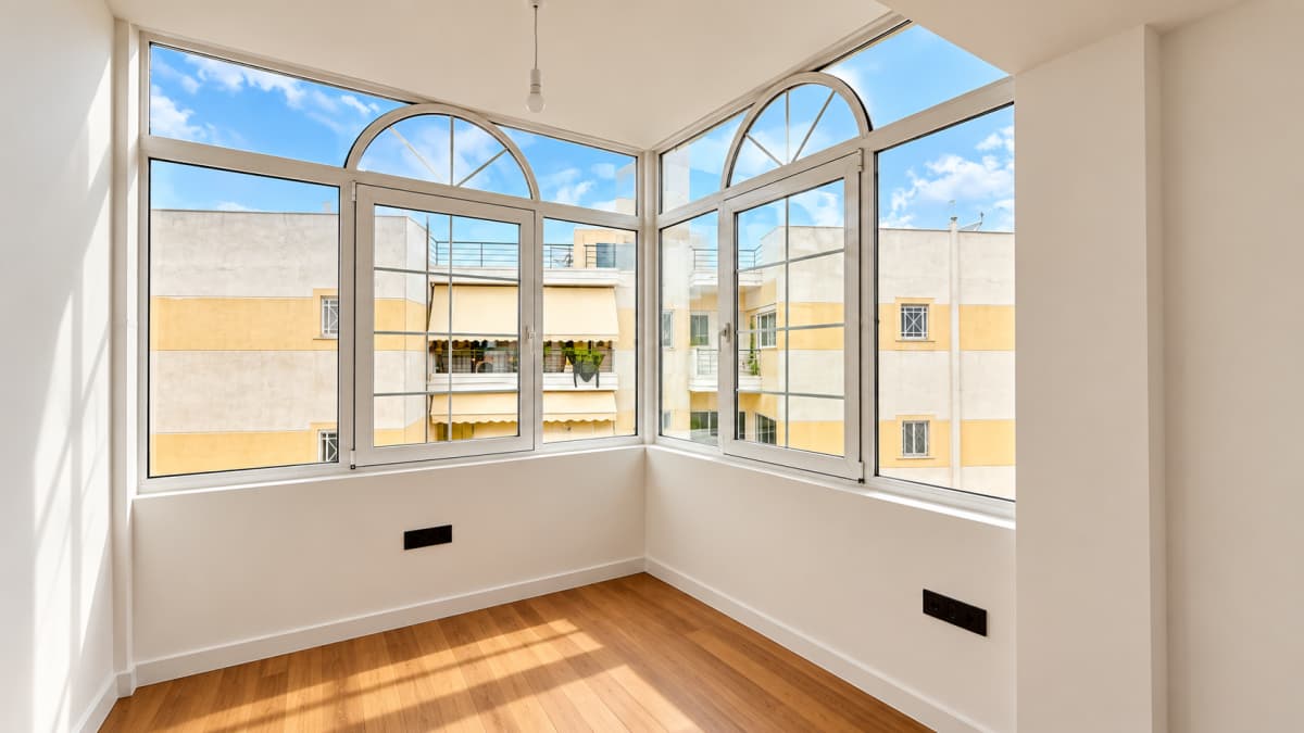 An empty room with white walls, wood-look flooring, and large arched windows providing an outdoor urban view.