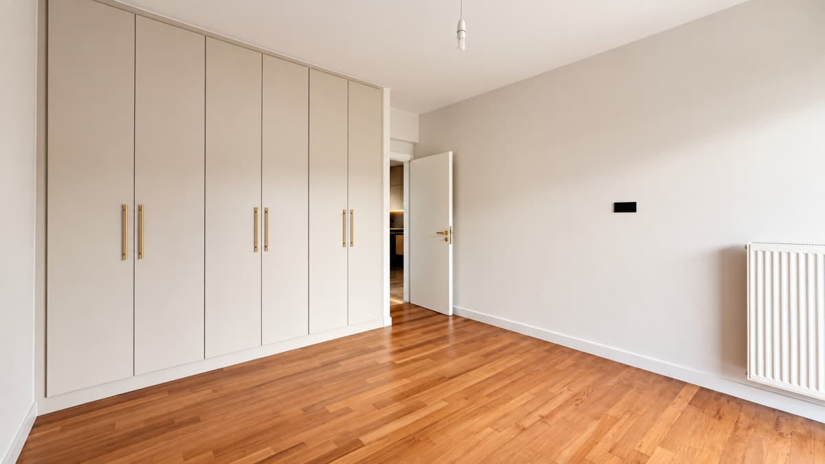 Empty room with a large built-in beige wardrobe on the left, wooden parquet flooring, white walls, and a radiator on the right wall.