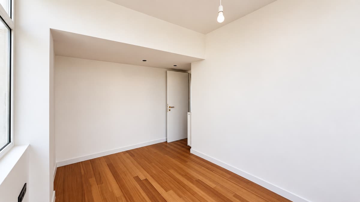 An empty room with white walls, a wooden floor, a single hanging light bulb, and a white door in the back.