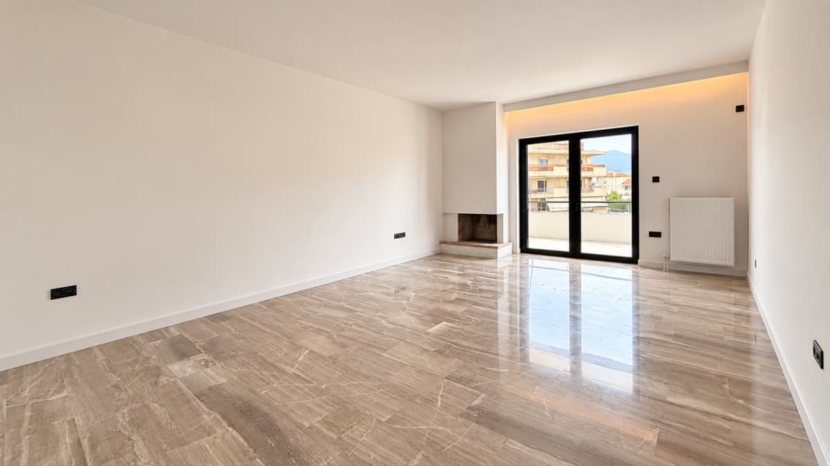Empty bright living room with white walls, polished brown marble floor, built-in fireplace, radiator, and sliding glass doors leading to a balcony.