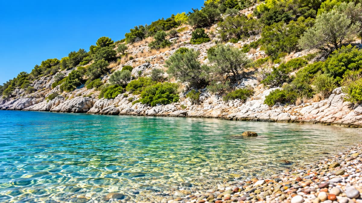 Clear turquoise sea water laps against a pebble beach beneath a steep, rocky, vegetated hillside under a blue sky.