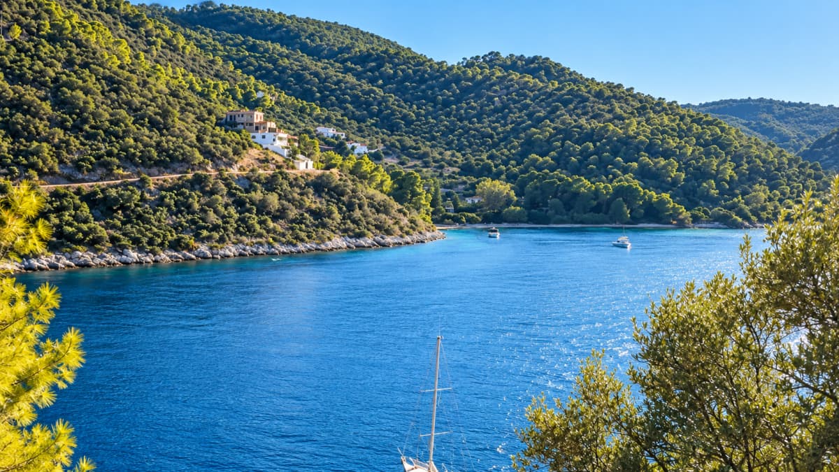 Wide view of a blue sea and bay surrounded by green hills, with white houses on the slope and a sailboat.
