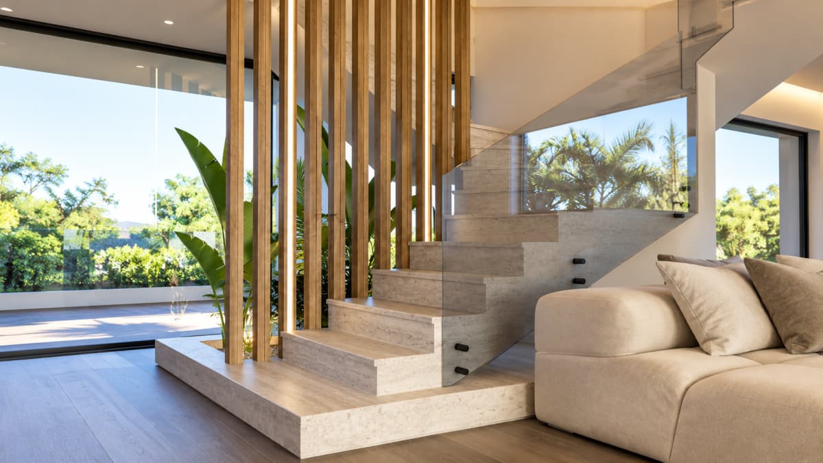Interior shot featuring a modern floating staircase with marble steps, a glass railing, wooden vertical slat partitions with integrated lighting, indoor plants, and a portion of a cream-colored sofa against a large window with outdoor scenery.