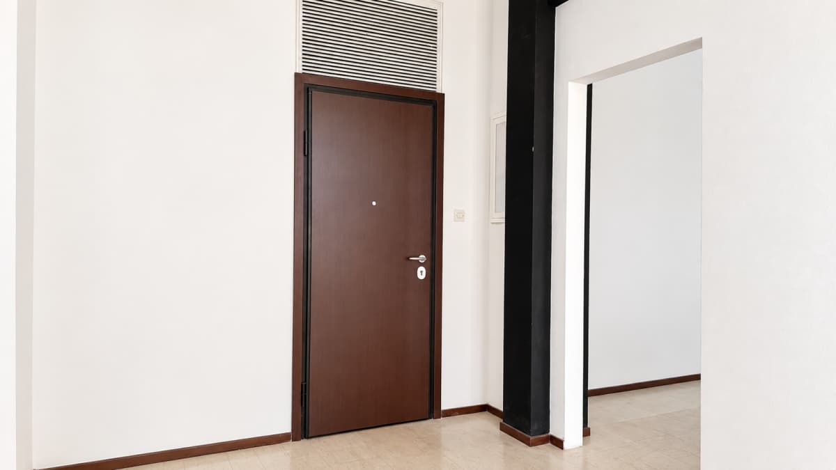 Λευκός διάδρομος με καφέ πόρτα, αεραγωγό από πάνω, μαύρη κολώνα και άνοιγμα προς άλλο δωμάτιο.