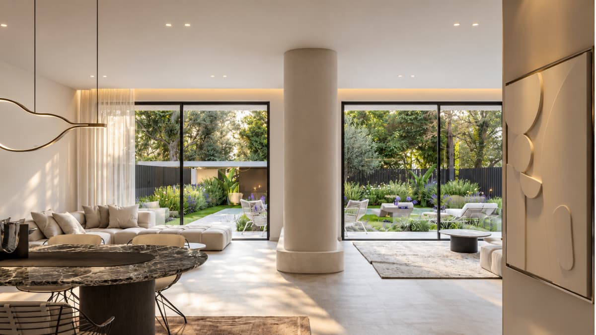 Interior living room with large glass sliding doors opening to a garden, a dining table in the foreground, and a central structural column.