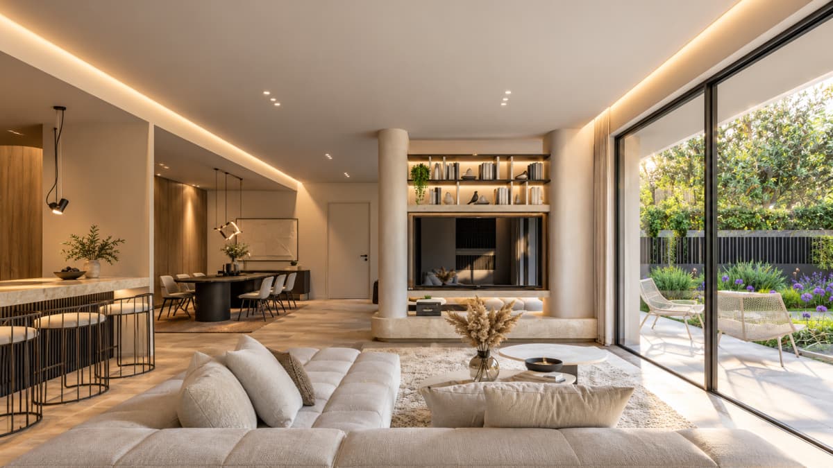 A modern open-plan living room with a large beige sectional sofa, a TV integrated into a wall unit between two columns, a dining area in the background, and large glass sliding doors looking out onto a garden patio.