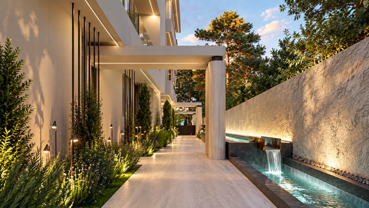 A modern exterior hallway with a long water feature, stone wall, cylindrical columns, and illuminated landscaping alongside a paved walkway.