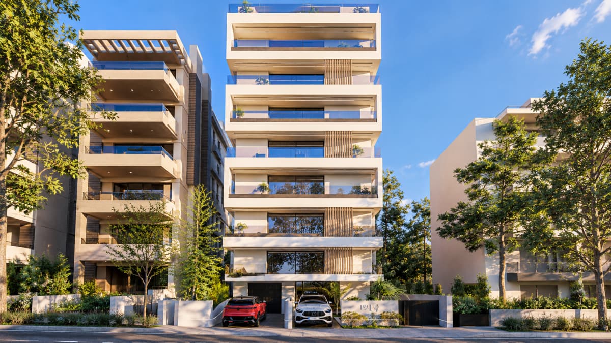 Street-level view of a modern multi-story residential building with balconies and parked cars, flanked by apartment buildings and trees.